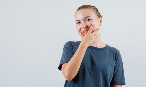 Young woman holding hand on her chin in grey t-shirt and looking cheery. front view.